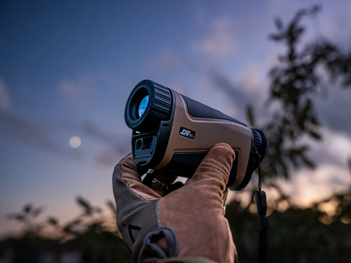 Gloved hand raising a DNT Hound thermal monocular against the dusk sky